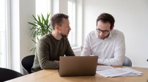 A compliance manager and an IT lead in business-casual clothing sit at a light oak table in a bright meeting room with tall windows, reviewing printed documentation and an open laptop showing indistinct interface shapes.