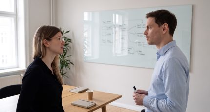 Two colleagues in business-casual clothing stand at a whiteboard in a bright meeting room with tall windows, working through a network access diagram together, with laptops and a coffee cup on the light oak table behind them.