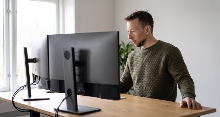 Two colleagues in business-casual clothing in a bright meeting room beside a tall window, one standing and pointing at a wall-mounted display showing indistinct dashboard shapes, the other seated at a light oak table with an open laptop.
