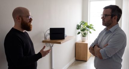 A network engineer in business-casual clothing stands at a standing desk in a bright open-plan office, reviewing a multi-site firewall inventory on two monitors, with a printed rack diagram on the desk and daylight from a floor-to-ceiling window.