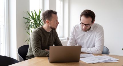 A compliance manager and an IT lead in business-casual clothing sit at a light oak table in a bright meeting room with tall windows, reviewing printed documentation and an open laptop showing indistinct interface shapes.