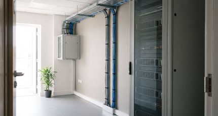 A network engineer in business-casual clothing stands in a bright equipment room checking labels on the front of a floor-standing comms cabinet, with a second engineer seated at a bench in the background and daylight from a window at the end of the room.