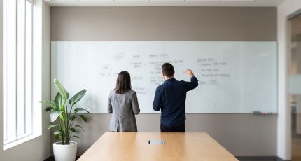 Three colleagues in business-casual clothing stand around a meeting-room table in early-afternoon daylight, one pointing at a printed project schedule while a laptop shows a network diagram, with a window and a plant behind them.