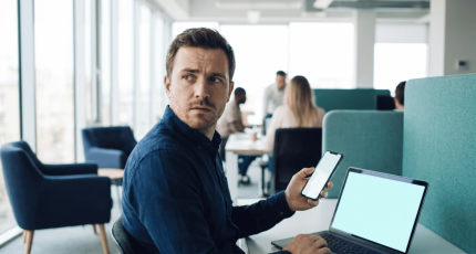 An office worker scanning their laptop screen with a personal phone, illustrating how quishing moves off the corporate network